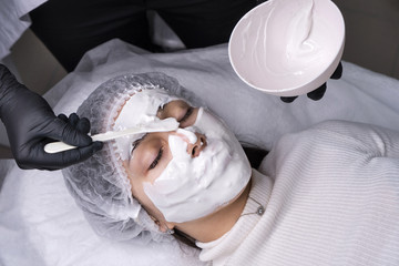 Fototapeta na wymiar Cosmetologist applies alginate mask with spatula on face of woman. Facial skin treatment in beauty salon. Anti-aging treatment. Cosmetology and professional facial skin care.