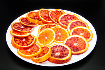 Naklejka na ściany i meble Sicilian sliced ​​orange on a white plate on a dark background. Sliced ripe Sicilian oranges against background. Healthy and tasty red sliced ​​orange.