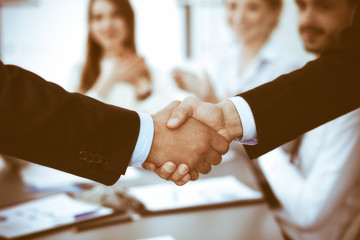 Business people shaking hands at meeting while theirs colleagues clapping and applauding. Group of unknown businessmen and women in modern white office. Success teamwork, partnership and handshake