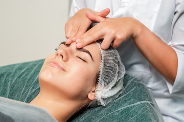 Fototapeta na wymiar Beautiful woman receiving forehead massage by hands of cosmetologist in beauty salon.