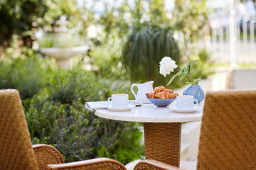 Morning coffee with fresh pastries and news in the newspaper in the fresh air on the terrace by the house. Plate with pastries, coffee and cream on a table decorated with a vase with a white rose