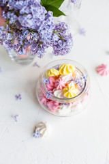 Some meringue kisses in a glass jar on the wait background decorated with bouquet of fresh lilac flowers. Spring season.