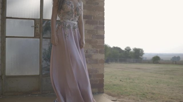 Senior high-school girl standing at old derelict buildings door with  blush prom dress blowing in the wind in slow motion