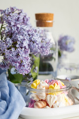 Some meringue kisses in a glass jar on the wait background decorated with bouquet of fresh lilac flowers. Spring season.