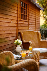 A table in the courtyard is served for Breakfast and decorated with a vase of flowers. On a wicker table there is orange juice, pastries and jam, and a fresh press