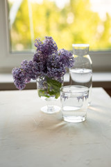 Fresh lilac flowers in a glass vase and fresh water in a glass are on the white table. Spring concept.