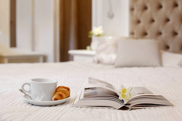 A Cup of coffee, a croissant and an open book lie on the bed in a bright cozy room, a delicate flower lies on the book.Time for a break. Space for text