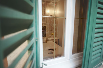 View through the half-open shutters and window on the interior of the bedroom. A Cup of coffee, a croissant, and an open book lie on the bed