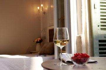 A bouquet in a vase, a glass of white wine and a plate with fresh raspberries and red currants are on the table in the room