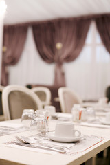 The table is served for Breakfast in a cozy bright dining room with large Windows and dark curtains. On the table are ceramic cups, glasses and Cutlery