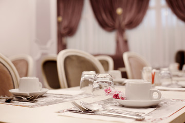 The table is served for Breakfast in a cozy bright dining room with large Windows and dark curtains. On the table are ceramic cups, glasses and Cutlery