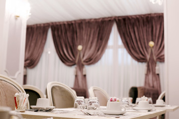 The table is served for Breakfast in a cozy bright dining room with large Windows and dark curtains. On the table are ceramic cups, glasses and Cutlery