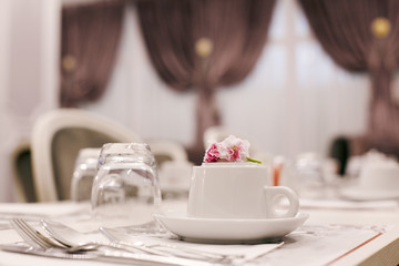 The table is served for Breakfast in a cozy bright dining room with large Windows and dark curtains. On the table are ceramic cups, glasses and Cutlery