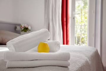 Beautifully and neatly folded white towels lie on the bed in the room against the background of the window. Towels are decorated with a delicious yellow Apple