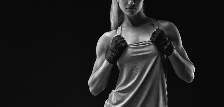 Black and white horizontal portrait of young woman with muscular body standing against black background. Image of fitness woman in sports clothing and gloves.