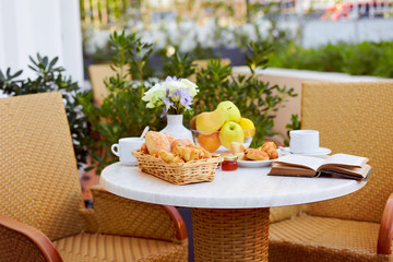 Fresh pastries, fruit, coffee, jam and an open book are placed on a table decorated with a bouquet of delicate flowers on the terrace. A romantic Breakfast is served in the open air