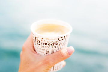A female hand is holding a cup of coffee in front of sea and blurred view 