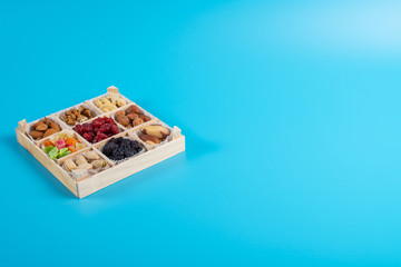 Assorted dried fruits and nuts in a wooden box isolated on a blue background. Raisins, candied fruits, cherries, Brazil nuts, hazelnuts, walnuts, almonds, cashews, pistachios.