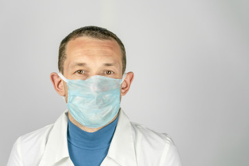 Male doctor in a medical gown and medical mask stands on a light background and looks at the camera with copy space.