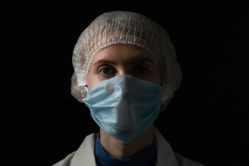 Young female doctor in medical mask, medical gown and cap on black background with copy space