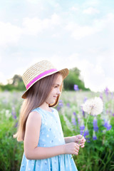 Beautiful little girl in a straw hat and dress holds a dandelion in a flowering field. Nature concept. A smiling girl is playing on the summer lawn. Allergy. Childhood. Copy space. Summer joy 