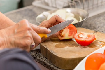 Old woman hands cutting a tomato on wooden board with knife in kitchen.