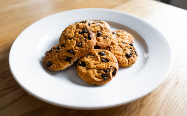 Choc chip cookies in a white plate on a wooden table. Close-up of homemade chocolate chip cookies. Basic drop cookies with chocolate morsels. Traditional handmade chewy cookies with chocolate chunk