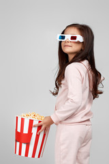 Obraz premium cute child girl wearing red-blue 3d glasses having fun and holding popcorn bucket isolated on gray background.