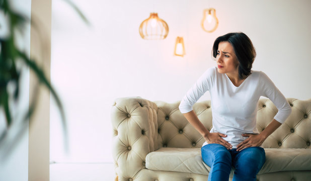 Menstrual disorder. Young woman is sitting on a sofa and touching her lower stomach while suffering from period pain.