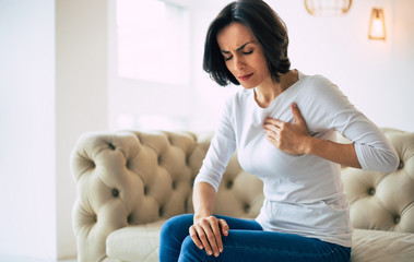Chest pain. Close-up photo of a brunette girl who is sitting on a couch with her eyes closed and her left hand touching her heart area.