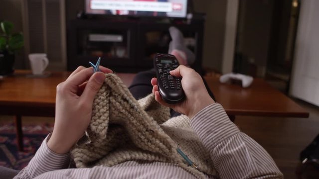 Woman changes the channel on the TV with a remote while crocheting on the couch