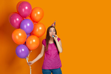 Fototapeta na wymiar happy funny young woman in pink t-shirt and birthday hat with bright colorful air balloons blowing party horn isolated on orange background. space for text