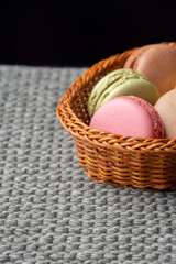 Top view of colored macarons in small basket, with selective focus, on gray knitted blanket, and black background, vertically, with copy space