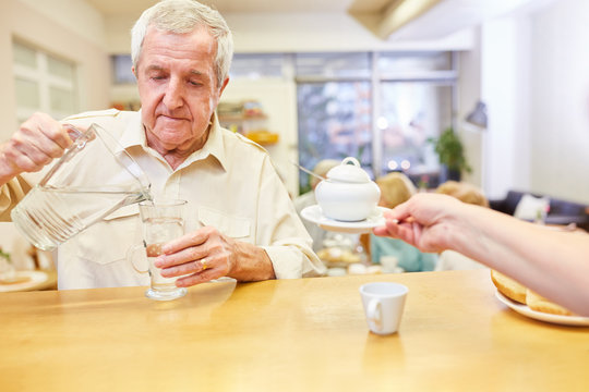 Senior gießt Wasser aus einer Karaffe in ein Glas im Aufenthaltsraum im Seniorenheim