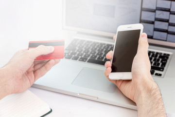 Male hands hold a phone and a bank card in front of a laptop monitor. Online earnings during the coronavirus pandemic. Close-up. Flare.