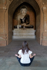 A female is meditating in lotus position, sitting on the floor in front of a Buddha statue in an ancient temple in Bagan in Myanmar.