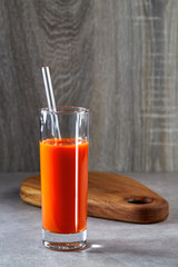 Glass of freshly squeezed carrot juice with a glass reusable tube stands on the concrete surface of the table against the background of a beautiful oak cutting Board.