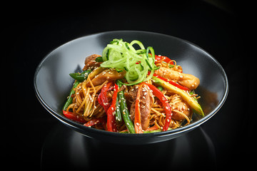 Appetizing wok egg noodles with chicken, bell pepper, asparagus, sesame seeds and fresh cucumber in a black bowl on a black background with reflection. Asian street food