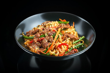 Appetizing wok udon noodles with beef, bell pepper, asparagus, sesame seeds in a black bowl on a...