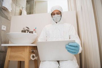 man in hazmat suit, medical mask, latex gloves and goggles using laptop in bathroom near washbasin