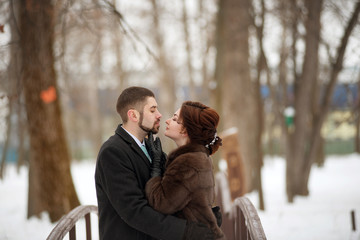A woman in a fur coat reaches out to kiss her bearded man in a winter park.