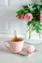 Spray drops splash in a pink cup of tea. Sunny morning with peonies flowers on a background. Beautiful breakfast idea. Self-isolation on quarantine.