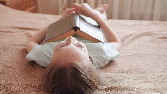 Laughing girl lying with book on bed. Cute smiling young girl looking aside while lying on bed with book. Leisure concept