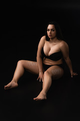 Fototapeta na wymiar Plus size model in underwear with lettering I Did not Ask For It on body sitting isolated on black