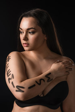 Portrait of plus size model in black bra with lettering hashtag feminism on body isolated on black