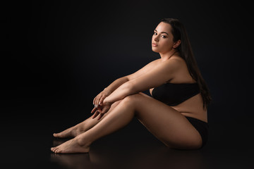Side view of beautiful plus size girl in underwear sitting on black background