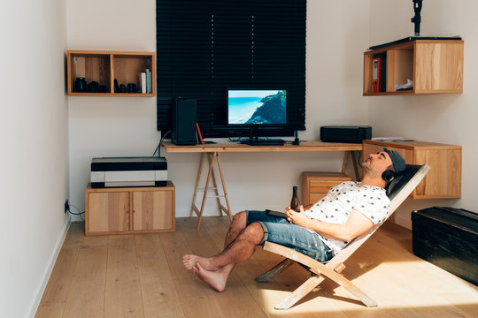 man with headphones and beer relaxing on deck chair at home with holiday pictures on the desktop screen - funny creative way of spending holidays and weekends in home isolation quarantine