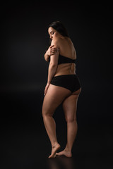 Fototapeta na wymiar Full length view of plus size girl hiding body with shame on black background