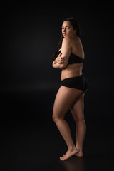 Fototapeta na wymiar Full length view of plus size girl hidding body with shame on black background