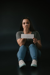 Naklejka premium Front view of plus size girl covering mouth with hand and holding empty card on black background